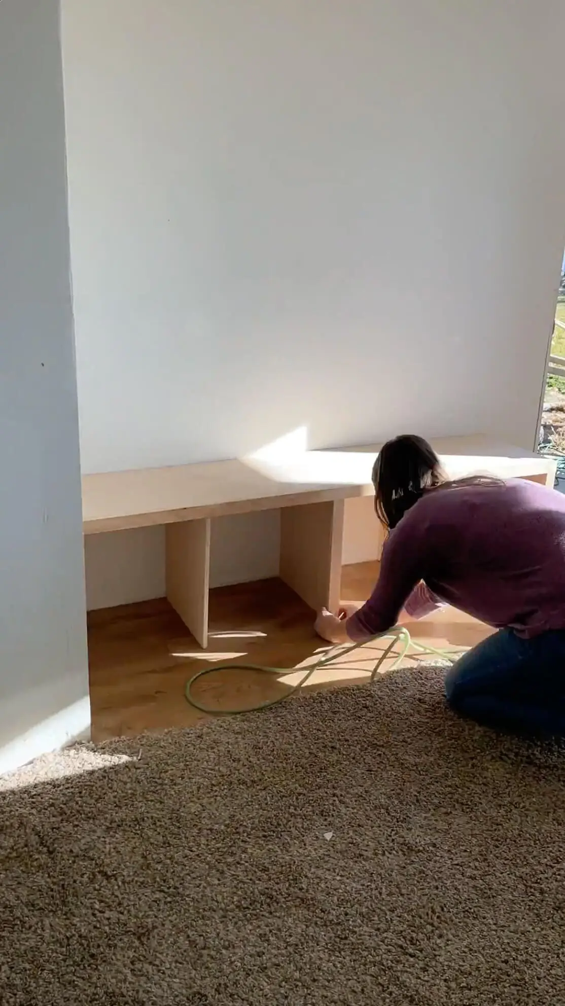 A woman kneeling on the floor, putting the finishing touches on a built-in mudroom storage bench with storage cubbies. The structure is nearly complete, positioned neatly against a white wall in a well-lit space with modern wood flooring.